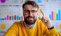 Caucasian male data analyst in yellow sweater pointing up with confident smile, against background of business charts and graphs. Professional workplace setting.