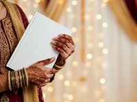 Bride in traditional wedding attire holding invitation card, displaying henna-decorated hands and gold bangles against warm bokeh lights backdrop.
