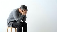 Middle-aged Asian man sitting on stool with head in hands, expressing stress, grief, or depression against light background.