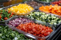 Fresh chopped vegetables and fruits in serving containers at salad bar, including tomatoes, cabbage, cucumber, mango, and carrots arranged in rows.