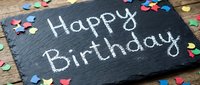 Happy Birthday written in chalk on a dark slate board surrounded by colorful confetti stars on a rustic wooden background.