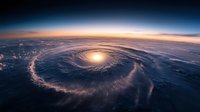 Aerial view of dramatic spiral storm system over ocean at sunset, with swirling clouds forming eye pattern against glowing horizon and dark atmosphere.