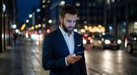 Young professional businessman using smartphone on city street at night with blurred traffic lights and urban bokeh background for business communication concepts.