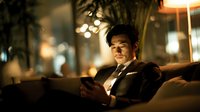Young Asian businessman in suit relaxing on sofa using smartphone in modern office lounge with warm evening lighting and city bokeh background.