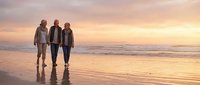 Three senior friends walking together on beach at sunset with golden sky and ocean waves reflecting on wet sand creating peaceful retirement moment.