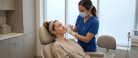 Professional dental hygienist in blue scrubs performing oral examination on relaxed female patient in modern clinic chair with natural lighting.