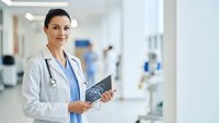 Professional female doctor holding medical tablet in modern hospital corridor with stethoscope around neck, smiling confidently at camera for healthcare concepts.
