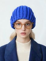 Young blonde woman wearing bright blue knitted beanie, transparent glasses, cream turtleneck and navy coat against light background. Fashion portrait in minimal style.