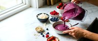 Hands pouring vibrant purple smoothie from blender into white bowl with fresh berries and oats on marble counter for healthy breakfast preparation.