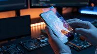 Hands holding smartphone displaying holographic circuit board interface in tech workspace with glowing monitors and computer equipment.