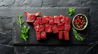 Raw beef cubes arranged in cow shape on dark slate board with herbs and peppercorns for culinary presentation and cooking preparation.