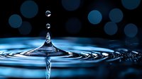 Clear water drop falling into calm blue surface creating symmetrical splash crown and ripples against dark background with bokeh lights. For wellness and nature concepts.
