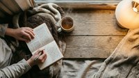 Cozy reading moment with hands holding open book, warm coffee cup and soft knitted blanket on rustic wooden surface creating peaceful atmosphere.