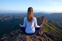 Young woman in white long sleeve top and purple pants meditating in lotus position on mountain peak at sunrise, overlooking scenic valley and mountain ranges.