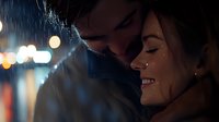 Young romantic couple in intimate moment during evening rain, with soft bokeh lights creating dreamy atmosphere. Woman smiling with closed eyes as man leans close.
