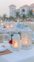 Elegant white lanterns with glowing candles on beach wedding table setting, featuring wine glasses and soft bokeh lights against resort background at dusk.
