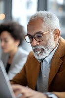 Senior African American businessman with glasses and white beard working on laptop in modern office environment.