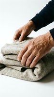 Senior man with weathered hands folding stacked beige knit sweaters against white background. Suitable for laundry, textile care, and domestic lifestyle content.