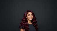 Attractive woman with long burgundy wavy hair and warm skin tone wearing black top against dark background. Suitable for beauty, hair care, and salon marketing materials.