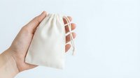 Hand holding a small natural cotton drawstring pouch against a light background, eco-friendly reusable bag for gifts or storage.