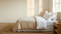 Minimalist bedroom interior with natural sunlight streaming through window onto unmade bed with neutral beige and white bedding on wooden floor.
