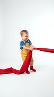 Happy toddler boy in yellow overalls playing with red fabric on white background. Joyful child development and creative play concept for parenting materials.