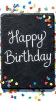 Happy Birthday written in chalk on a dark slate board surrounded by colorful confetti on white background, festive celebration concept.