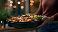 Grilled meat and vegetables served in cast iron pan with bokeh lights background, fresh herbs garnish, hands holding hot dish for dinner party.