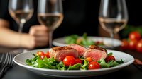 Medium rare sliced beef steak served on white plate with fresh arugula salad, cherry tomatoes and wine glasses in background, restaurant setting.