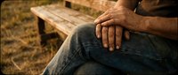 Senior man in denim jeans sitting on wooden park bench outdoors during golden hour sunset. Elderly hands resting on knee showing peaceful retirement moment.