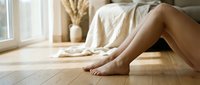 Bare legs of a young woman relaxing on a warm wooden floor at home, soft natural light, cozy bedroom interior in background.