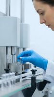 Female scientist in white lab coat and blue latex gloves operating laboratory equipment, conducting research or analysis in sterile medical or pharmaceutical facility environment.