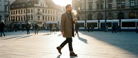 Young businessman walking confidently across sunny European city square during golden hour with historic architecture and modern tram in background.