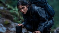 Young Asian woman in black raincoat and backpack crouching near portable speaker in rainy forest setting, creating atmospheric outdoor adventure scene.