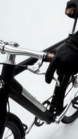 Close-up of a gloved hand gripping the brake lever of a sleek black electric folding bike with silver handlebars.