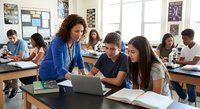 Female teacher with curly hair in blue blazer helping diverse high school students with laptop computer in modern classroom setting.