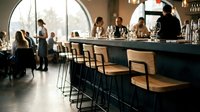 Modern bar counter with wooden woven seats and metal frames, bartender serving drinks to guests in contemporary lounge with large windows and warm lighting.