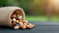Mixed nuts spilling from rustic burlap sack onto dark wooden surface against blurred green garden background, natural healthy snack food composition.