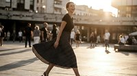 Young woman with fair complexion wearing a flowing black polka dot dress, dancing in an urban plaza during golden hour with blurred pedestrians and architecture in background.