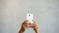 Hands of a middle-aged person with medium brown skin tone holding a white smartphone with camera visible, against neutral gray background for technology and communication concepts.