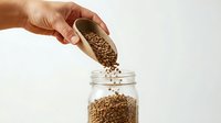 Caucasian hand pouring lentils from wooden scoop into glass jar on white background, healthy eating and food storage concept.