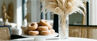 Fresh glazed donuts on white plate in modern kitchen with pampas grass decoration and natural lighting for bakery and breakfast concepts.