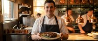 Male chef with dark hair and light skin, wearing glasses, white shirt and black apron, holding plated gourmet dish in modern restaurant kitchen with copper pendant lights and blurred staff background.