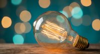Vintage Edison light bulb with glowing filament on wooden surface against colorful bokeh background. Retro lighting concept for creative projects and designs.