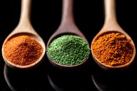 Vibrant spice powders in wooden spoons against black background - orange paprika, green matcha tea and turmeric curry powder arranged in row, macro food photography.