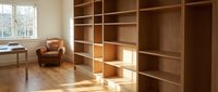 Modern minimalist home office with built-in wooden shelving unit and leather armchair by window. Clean contemporary interior design for workspace organization.