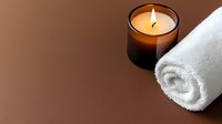 Minimalist spa composition with white rolled towel and aromatic candle in dark glass holder on brown background. Wellness and relaxation concept.