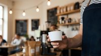 Barista holding white takeaway cup in modern cafe with blurred customers and wooden shelves in background, suitable for coffee shop, hospitality and small business concepts.