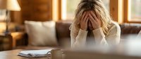 Stressed blonde woman covering face with hands sitting at wooden table in cozy home interior expressing overwhelm and mental fatigue.