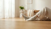 Cozy home relaxation scene with woman in knit socks sitting on wooden floor with soft blanket, natural light streaming through white curtains creating peaceful atmosphere.